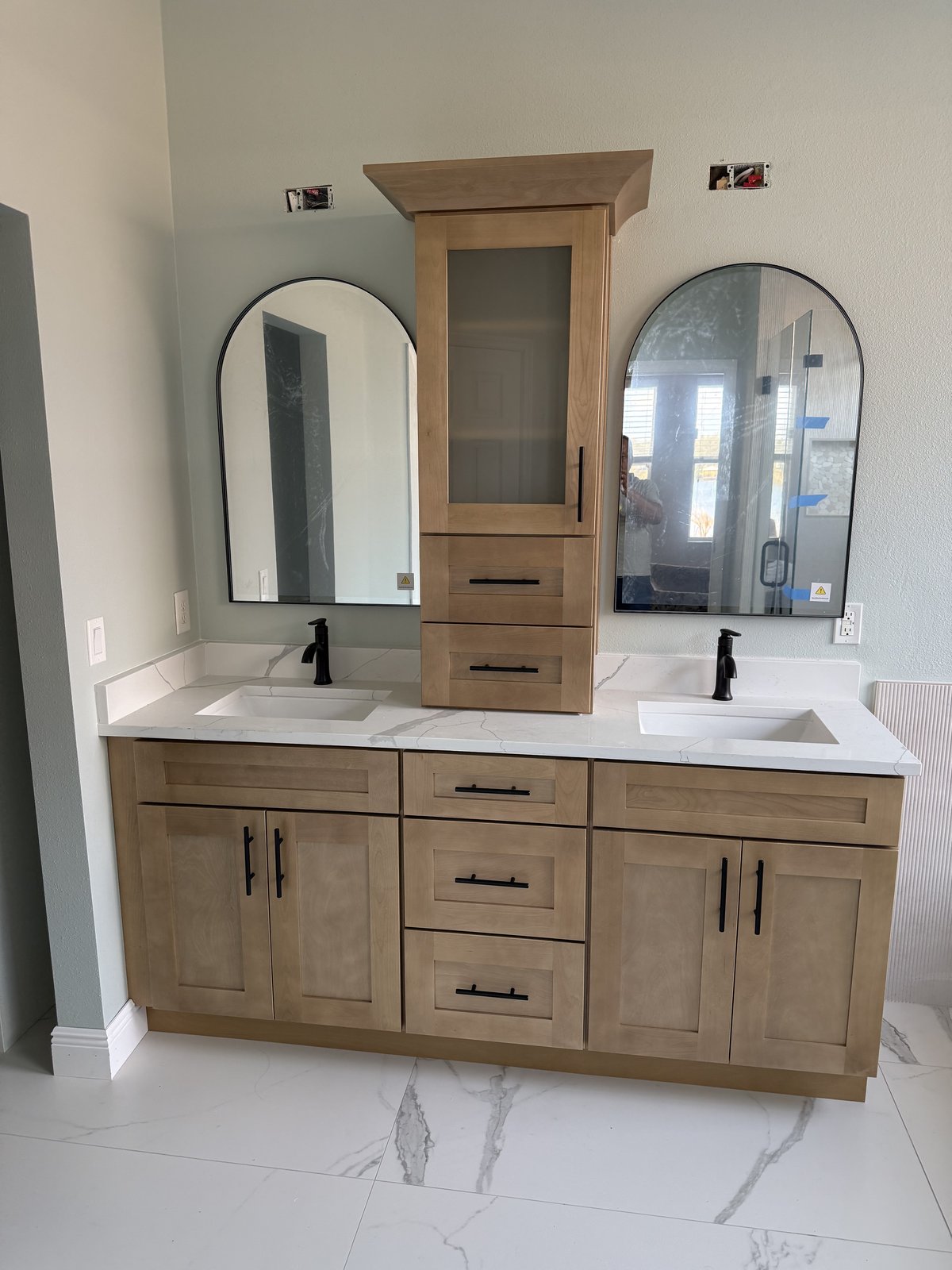Tocador doble de madera cálida con espejos arqueados, cubierta de cuarzo y grifería negra mate sobre relucientes pisos de porcelanato tipo mármol.