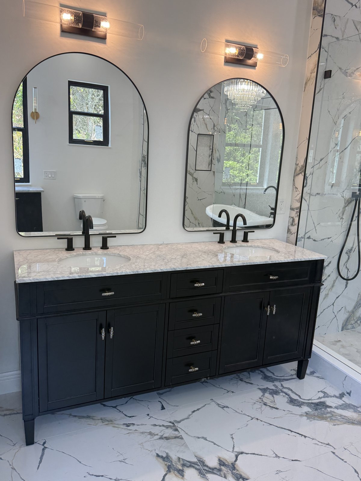 Elegante tocador doble en negro con cubierta de mármol Carrara, espejos arqueados y porcelanato tipo mármol en todo el espacio.