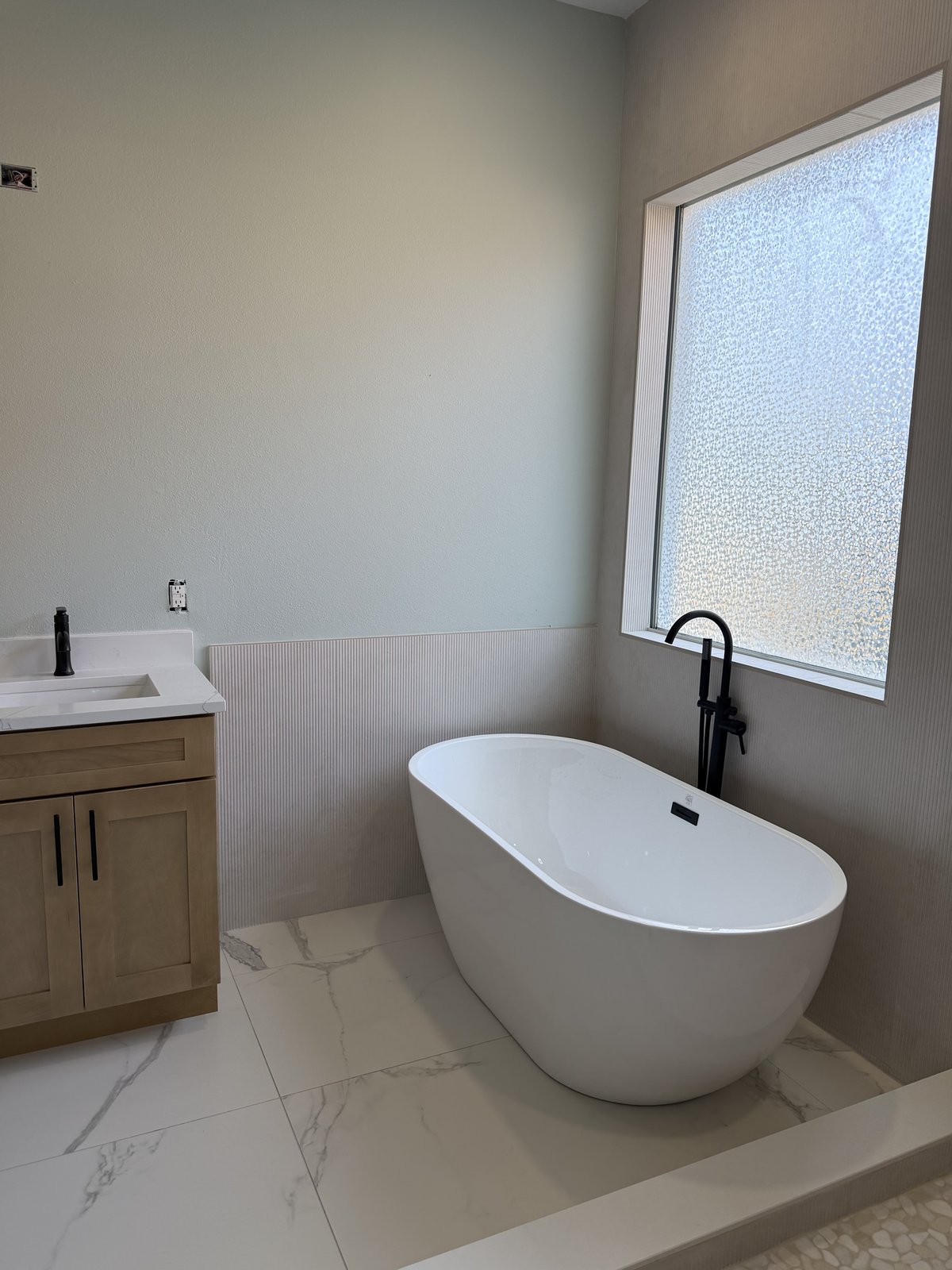 Tina exenta enmarcada por paneles de pared acanalados y vidrio esmerilado, rematada con pisos de porcelanato tipo mármol.