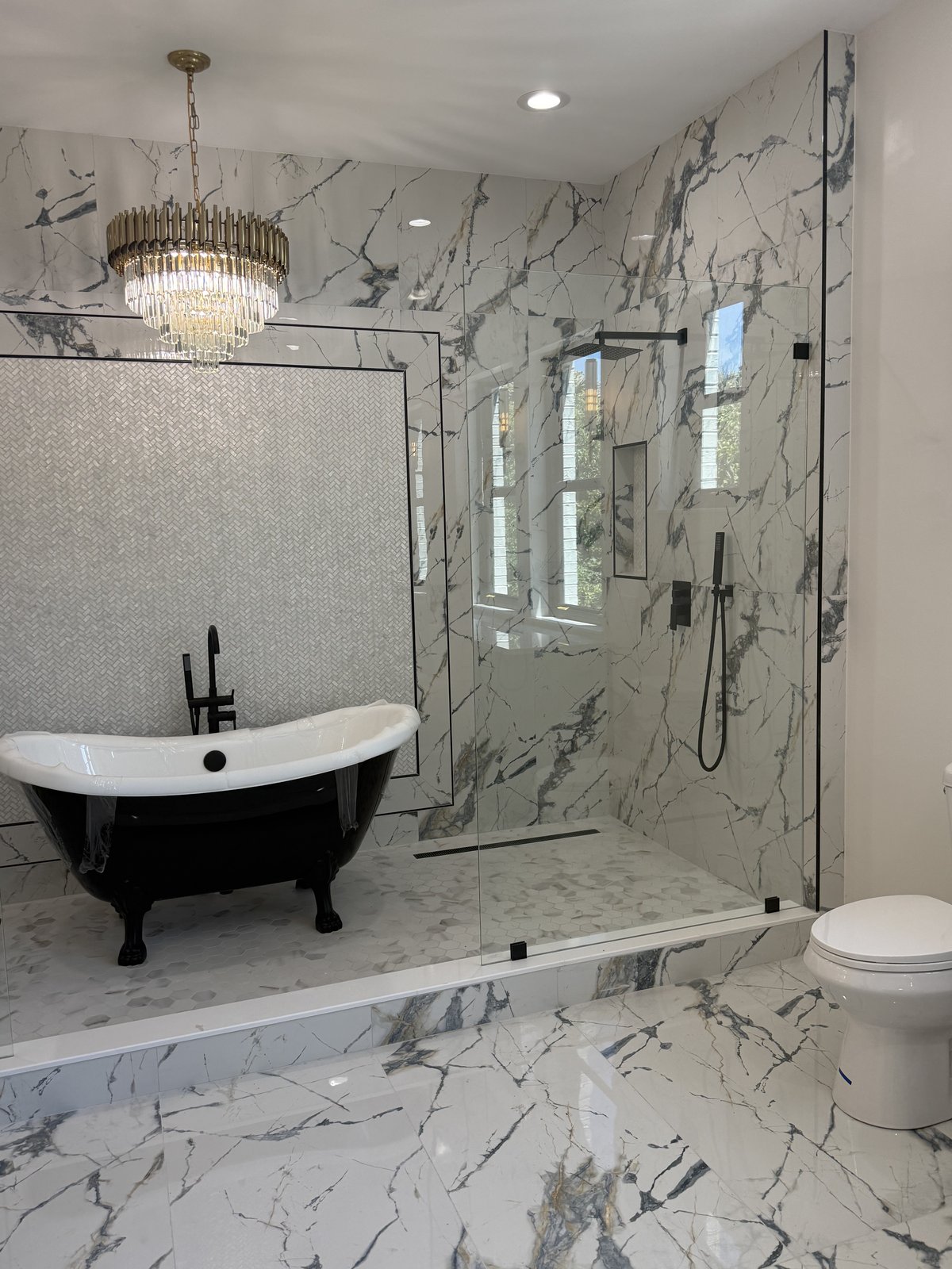 Pieza central de un baño principal de lujo: una tina clásica negra enmarcada por mosaico en espiga y un candelabro de cristal.